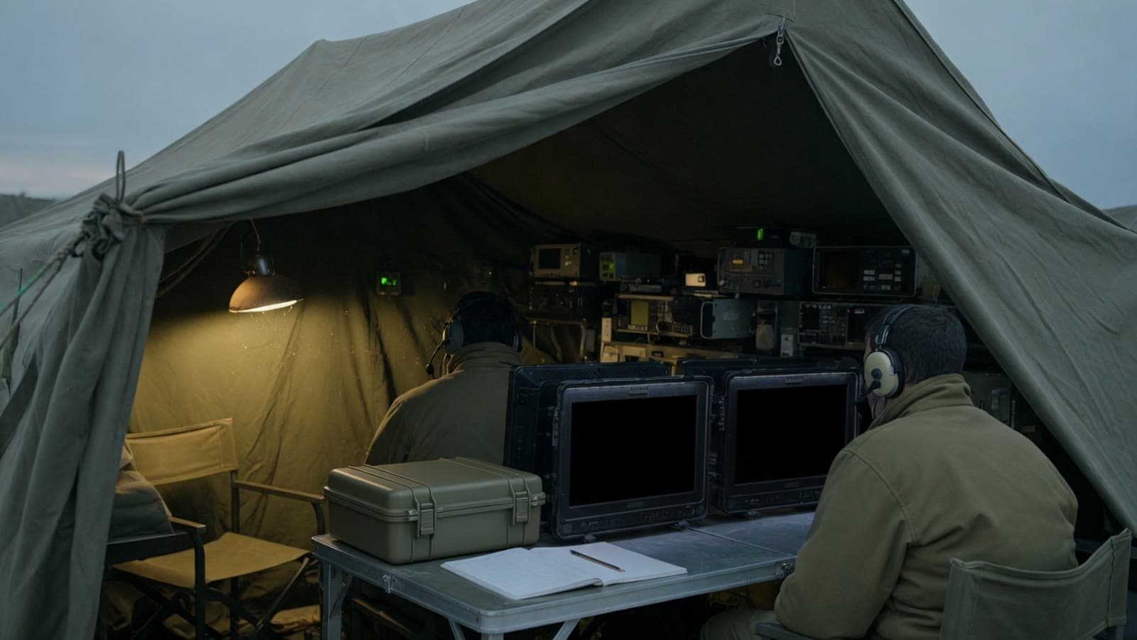 Two operators seen from the entrance of a field tent, with dark monitors, a plain case, and a blank log on the table. Conceptual.